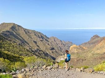 Hiker with blue backpack on rocky trail in Tenerife mountains. Steep gorge and ocean visible in background under clear blue sky.