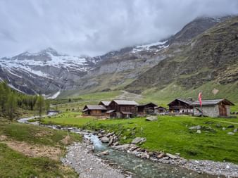 Mountain stream flowing through green meadows past Lazins Alm wooden chalets, with snow-capped peaks and cloudy sky in the background.