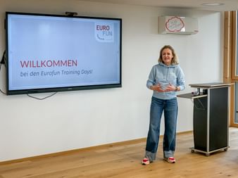Woman in blue hoodie standing in training room next to screen displaying 'WILLKOMMEN bei den Eurofun Training Days' with Eurofun logo.