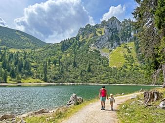 Erwachsener und Kind auf Uferweg am Spitzingsee mit Bergen und Wald. Blauer Himmel mit weißen Wolken über felsigen Gipfeln.