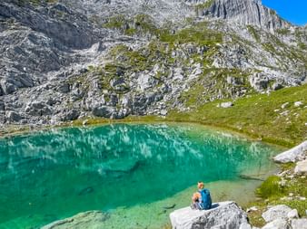 Hiker with backpack sitting on rock beside crystal-clear turquoise mountain lake in Allgäu Alps, surrounded by dramatic limestone cliffs.