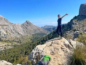 Hiker Julia stands on a rocky outcrop with raised arm on the GR221 trail in Mallorca, surrounded by dramatic mountain peaks and valleys.