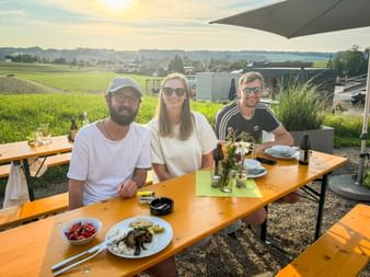 Three colleagues sitting at wooden picnic table with food and drinks, smiling at camera. Rural landscape with green fields visible in background.