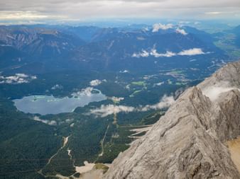 Luftaufnahme von der Zugspitze mit felsigen Berghängen, blauem See, bewaldeten Tälern und fernen Bergketten unter bewölktem Himmel.