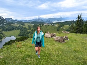 Wanderin in türkiser Jacke auf grüner Almwiese mit ruhenden Kühen, Bergsee und österreichischen Alpen im Hintergrund.