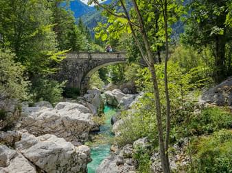 Stone arch bridge spanning turquoise Soča River in Julian Alps. Rocky riverbed with white boulders, lush green forest, and mountains in background.