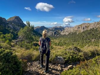 Hiker with backpack standing on rocky trail overlooking Tramuntana mountain range with green valleys and blue sky with white clouds.