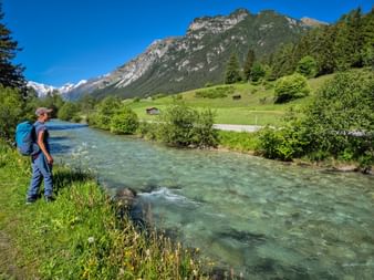 Wanderin mit blauem Rucksack am türkisfarbenen Gschnitzbach im Gschnitztal, mit grünen Wiesen, Bergen und blauem Himmel.