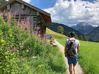 Wanderer mit Rucksack auf Schotterweg an Holzalmhütte mit rosa Blumen vorbei. Berge und grüne Wiesen unter blauem Himmel sichtbar.
