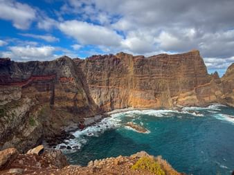 Towering layered rock cliffs at São Lourenço peninsula in Madeira meet turquoise ocean waters under a partly cloudy sky.
