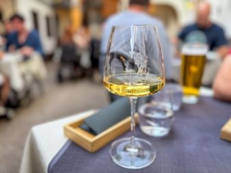 Glass of white wine on a restaurant table with blurred diners in the background. A wooden tray with napkin and water glass are nearby.