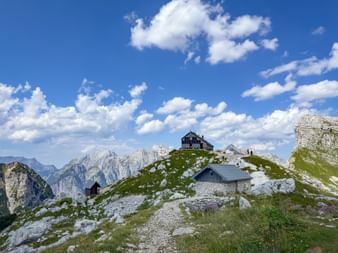 Mountain huts on a rocky ridge in the Julian Alps with jagged peaks in the background under a blue sky with white clouds.