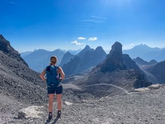 Wanderin mit blauem Rucksack steht auf felsigem Gelände und blickt auf dramatische Berggipfel der Allgäuer Alpen unter klarem blauen Himmel.
