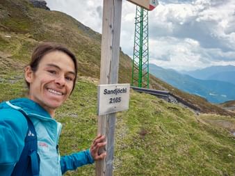 Lächelnde Frau in türkisfarbener Jacke am Sandjöchl-Gipfelkreuz (2165m). Berghänge und entfernte Gipfel unter bewölktem Himmel sichtbar.