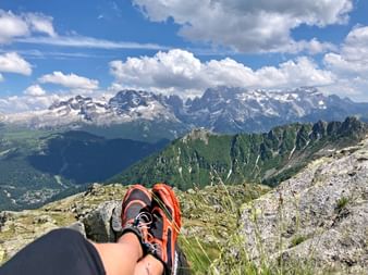 Hiker's feet in orange hiking boots resting on rocky summit with panoramic view of Brenta Dolomites mountain range under blue sky with clouds.