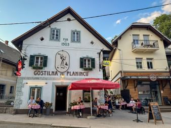 White traditional Slovenian guesthouse Gostilna Pri Planincu from 1903 with outdoor seating under red umbrellas and guests dining.