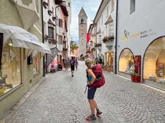 Wanderer mit rotem Rucksack in der Sterzinger Fußgängerzone. Der historische Zwölferturm erhebt sich im Hintergrund. Kopfsteinpflaster gesäumt von Geschäften.
