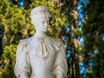 White marble statue of a woman in 19th-century dress at Achilleion Palace, with elaborate hairstyle and detailed clothing against green foliage.