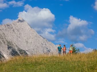 Drei Wanderer mit Rucksäcken stehen auf grasigem Hang im Leutascher Gaistal vor dramatischen Kalksteingipfeln und blauem Himmel.