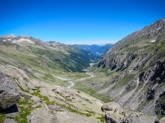 Weite Talansicht des Obersulzbachtals im Nationalpark Hohe Tauern mit steilen Felswänden, einem Gletscherbach und fernen Gipfeln unter blauem Himmel.