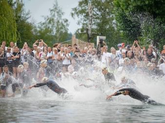 Mass swimming start at Trumer Triathlon with athletes diving into the water creating large splashes while spectators watch from the shore.