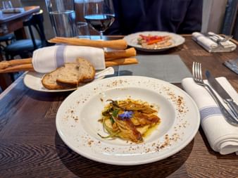 Braised fennel with caramelized onions on white plate, garnished with purple flower. Bread, wine glasses, and grissini on wooden table.