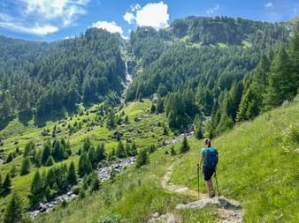 Hiker with blue backpack on mountain trail near Boffalora, Via Spluga. Green alpine meadows, dense forests, and mountain stream in valley below.