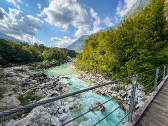 Holzsteg mit Metallgeländer über den türkisfarbenen Soča-Fluss bei Trnovo ob Soči in den Julischen Alpen, umgeben von grünen Wäldern und Bergen.