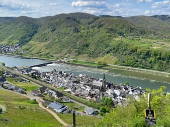 Panoramablick auf die Mosel, die sich durch grünes Tal mit Dorf, Kirche und Weinbergen schlängelt. Hügelige Landschaft und blauer Himmel.
