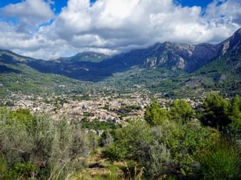 Panoramic view of Sóller town nestled in a valley, surrounded by the Tramuntana Mountains under a partly cloudy sky.