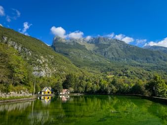 Plužna-Stausee mit grünem Wasser, das Berge und Häuser spiegelt. Bewaldete Gipfel der Julischen Alpen unter blauem Himmel mit weißen Wolken.