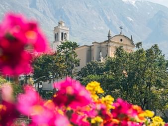 San Stefano Church in Malcesine with bell tower and dome, framed by pink and yellow flowers. Mountains visible in the background.