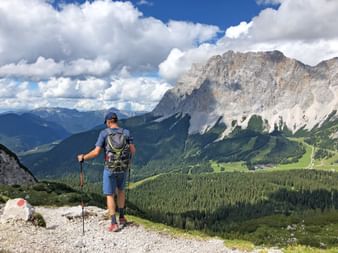 Wanderer mit Rucksack und Wanderstöcken auf felsigem Weg mit Blick auf dramatische Kalksteingipfel der Zugspitze-Region und grüne Täler.