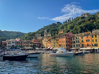 Boats moored in Portofino harbor with colorful buildings along the waterfront and green hills rising behind under blue sky.