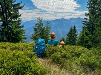 Hiker with blue backpack sitting on grassy hillside overlooking mountain panorama. Conifer trees frame the view of distant peaks under cloudy sky.