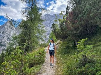 Wanderin auf Schotterweg in den Julischen Alpen am Vršič Pass, umgeben von Nadelbäumen mit felsigen Berggipfeln im Hintergrund.