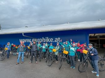Group of cyclists with yellow bags standing with bikes in front of blue Eurobike building under cloudy sky during Danube cycling tour 2025.