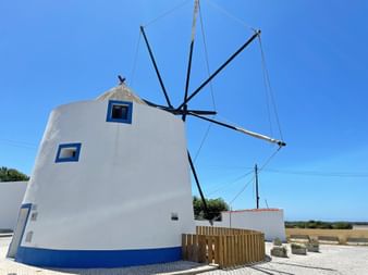 Weiße zylindrische Windmühle mit blauem Rand und Fenstern in Longueira. Schwarze Holzflügel ragen von der Struktur gegen klaren blauen Himmel.