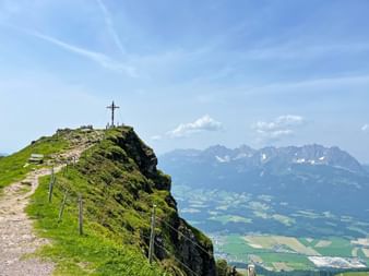 Summit cross on Kitzbüheler Horn with hiking path, green slopes, and mountain range in background. Valley with fields visible below under blue sky.