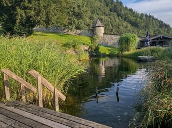 Natural swimming pond with wooden dock in foreground, Schloss Kammer castle with round tower on hillside, surrounded by forest in Pinzgau.