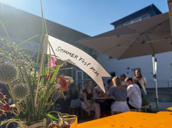 Blumenarrangement mit rosa Blüten und einem Schild mit der Aufschrift 'Sommer Fest 2026' auf einer Terrasse. Im Hintergrund sitzen Menschen unter Sonnenschirmen.