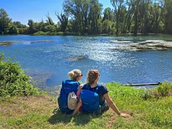 Two hikers with blue backpacks sitting on grassy riverbank overlooking the Sile River with trees and blue sky in background.