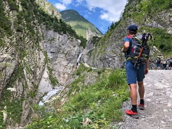 Wanderer mit Rucksack auf Schotterweg mit Blick auf dramatische Bergschlucht mit Wasserfall am Lechweg. Felsige Klippen und grüne Gipfel unter blauem Himmel.