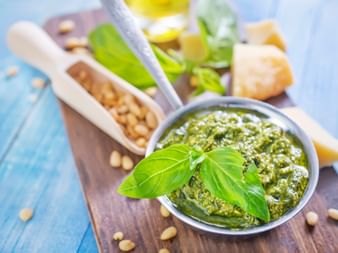 Bowl of green Pesto Genovese garnished with fresh basil leaves, surrounded by pine nuts, olive oil, parmesan cheese on a wooden board.