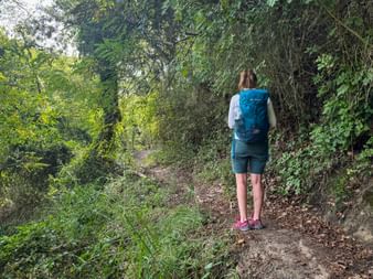 Wanderin mit blauem Rucksack auf schattigem Waldweg in der Toskana, umgeben von dichter grüner Vegetation und überhängenden Bäumen.