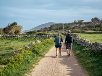Zwei Wanderer auf einem Feldweg zwischen Steinmauern bei Oia. Grüne Felder, Bäume und Berge im Hintergrund unter bewölktem Himmel.