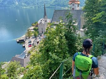 Hiker with green backpack descending path overlooking Hallstatt village with church spire, lakeside buildings, and blue alpine lake.