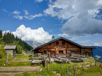 Traditional wooden alpine hut with picnic tables in Pinzgauer Grasberge, surrounded by green meadows, forests, and mountain peaks under blue sky.
