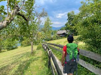 Hiker with green backpack walking on fenced path through meadow in Salzkammergut, with lake view, traditional house, and mountains under blue sky.