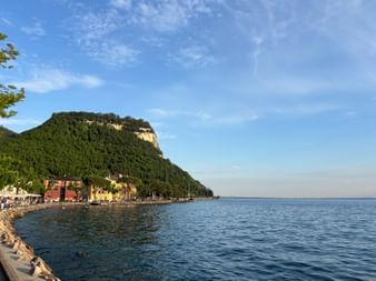 Uferpromenade in Garda mit bunten Gebäuden am Gardasee. Ein bewaldeter Berg erhebt sich hinter der Stadt unter blauem Himmel.